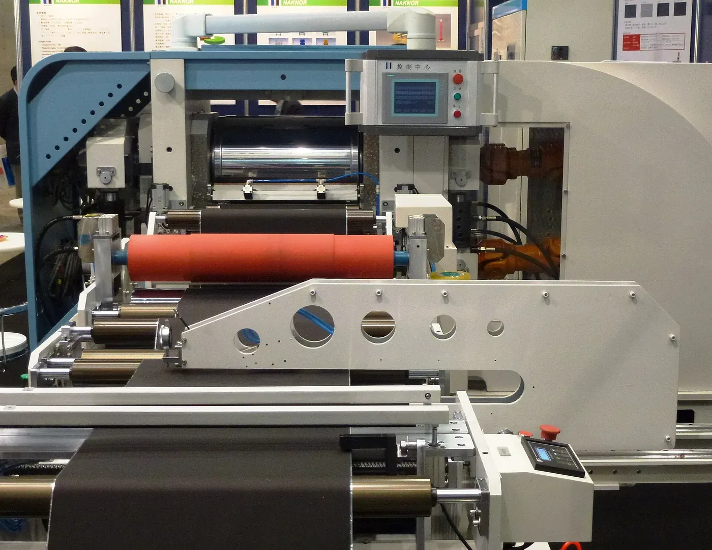 An industrial calendering machine with a dark, thin electrode film threaded between polished metal and red rubber rollers, control panel and hydraulic fittings visible behind it.