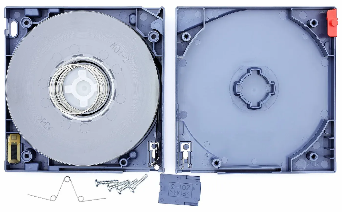 An opened data-tape cartridge shown from above, its reel of thin metallic-grey magnetic tape wound around a central hub, beside the cartridge's disassembled outer casing and a small pile of screws.