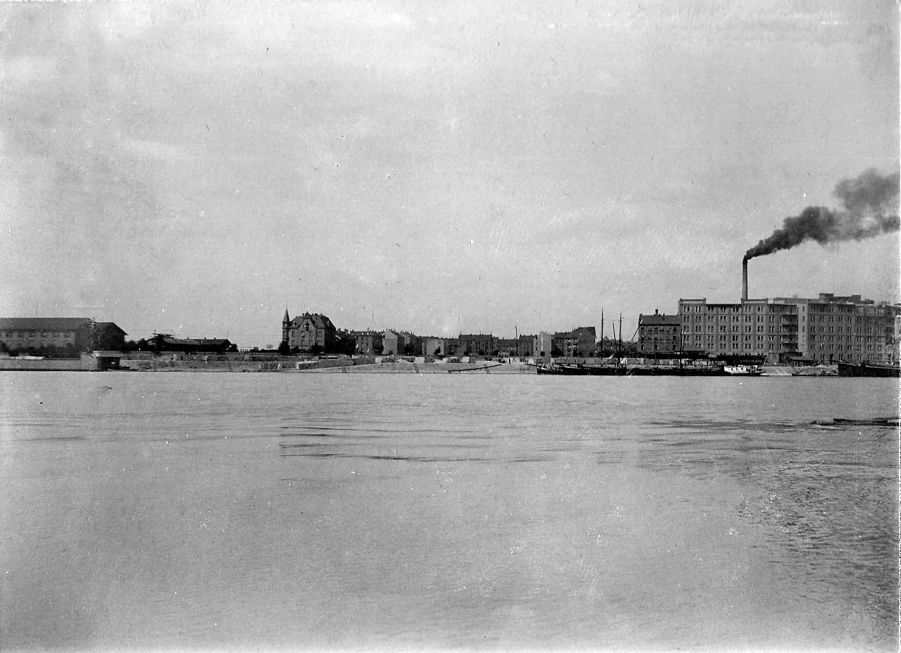 A wide river with the industrial waterfront of a German city on the far bank, warehouses and tenement blocks along the quay and a tall factory chimney trailing dark smoke.