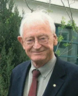 A smiling elderly man with white hair and glasses, wearing a dark blazer and a maroon tie, standing outdoors in front of garden greenery.