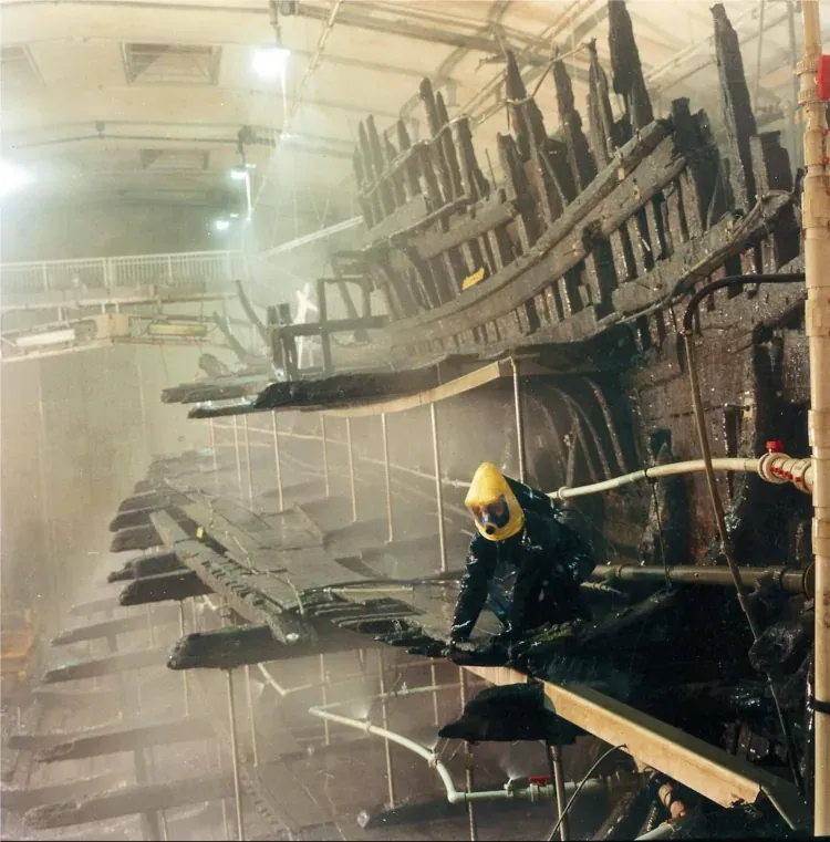 A conservator in protective coveralls and a yellow hood sprays a fine mist over the exposed, blackened timber ribs of a large wooden ship hull inside a purpose-built conservation hall.