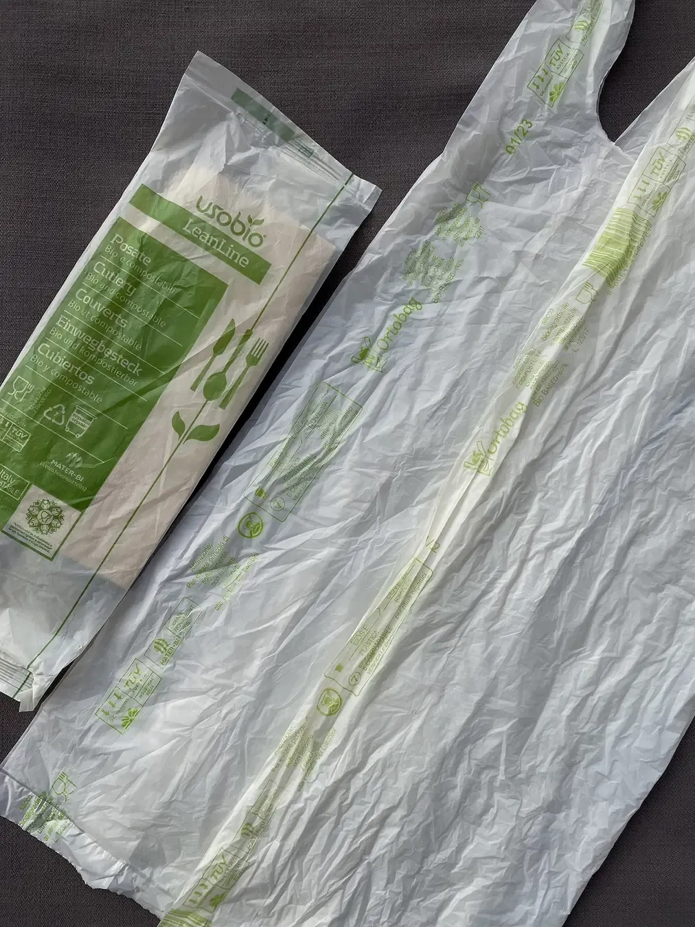 A folded compostable plastic bag printed with green leaf logos and multilingual 'bio and compostable' text, laid beside a wrapped set of disposable cutlery marked with the same certification symbols and the brand name Mater-Bi.