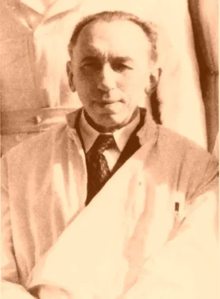 A sepia-toned portrait photograph of an older man with a receding hairline, wearing a white laboratory coat over a shirt and dark tie, a document rolled under one arm.