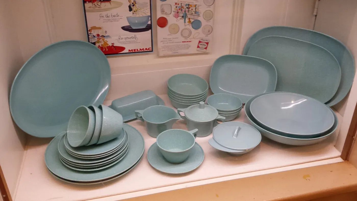A museum display shelf of pale blue Melmac dinnerware (plates, bowls, cups, a creamer and a covered butter dish) with vintage Melmac advertisements pinned to the wall behind.