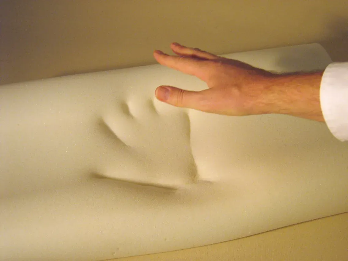 A hand pressing into a pale, off-white block of foam, which shows only a shallow, fading impression around the fingers as it springs back.
