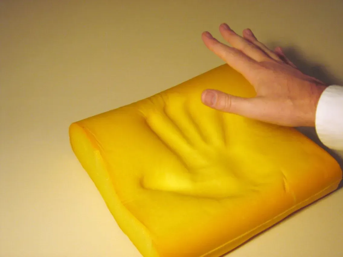 A hand hovering just above a bright yellow block of foam that retains a deep, detailed impression of fingers and palm after the hand has lifted away.