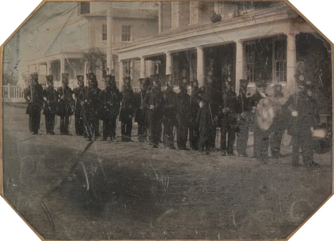 A daguerreotype of a company of volunteer soldiers in dark uniforms and tall shakos, muskets shouldered, standing in formation on a street in front of a colonnaded building, with a drummer at the rear of the line.