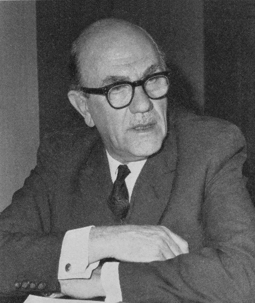 A balding man in glasses and a suit, photographed mid-conversation with one arm resting on a table.