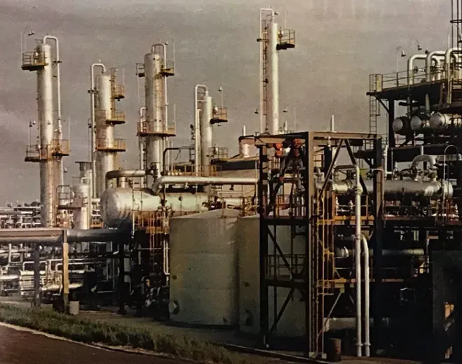 A colour photograph of an industrial petrochemical plant with tall distillation columns, storage tanks and pipework, photographed from ground level.