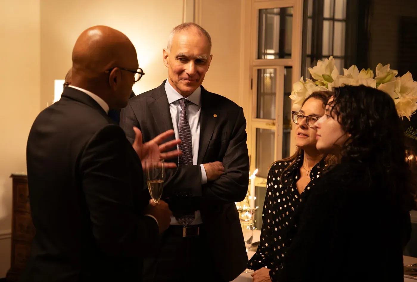 A group of people in formal evening dress talking at a reception; the central figure, an older man with grey hair wearing a Nobel medal pin on his lapel, gestures with one hand while holding a champagne glass in the other.