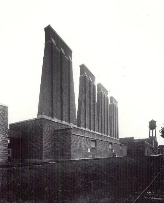 A brick paint and varnish factory with four tall, narrow chimney stacks of varying heights standing above the main building, a water tower visible behind.