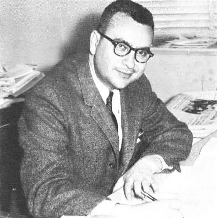 A black-and-white studio-style portrait of a man in a tweed jacket and glasses, seated at a desk with papers, smiling toward the camera.