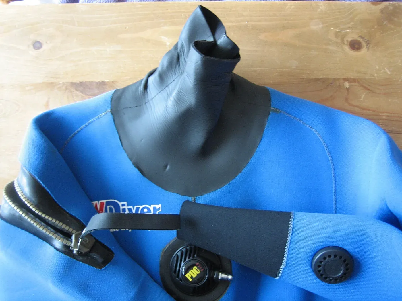 A close-up of a blue diving drysuit with black neoprene neck and wrist seals, a valve and a zip visible against a wooden background.