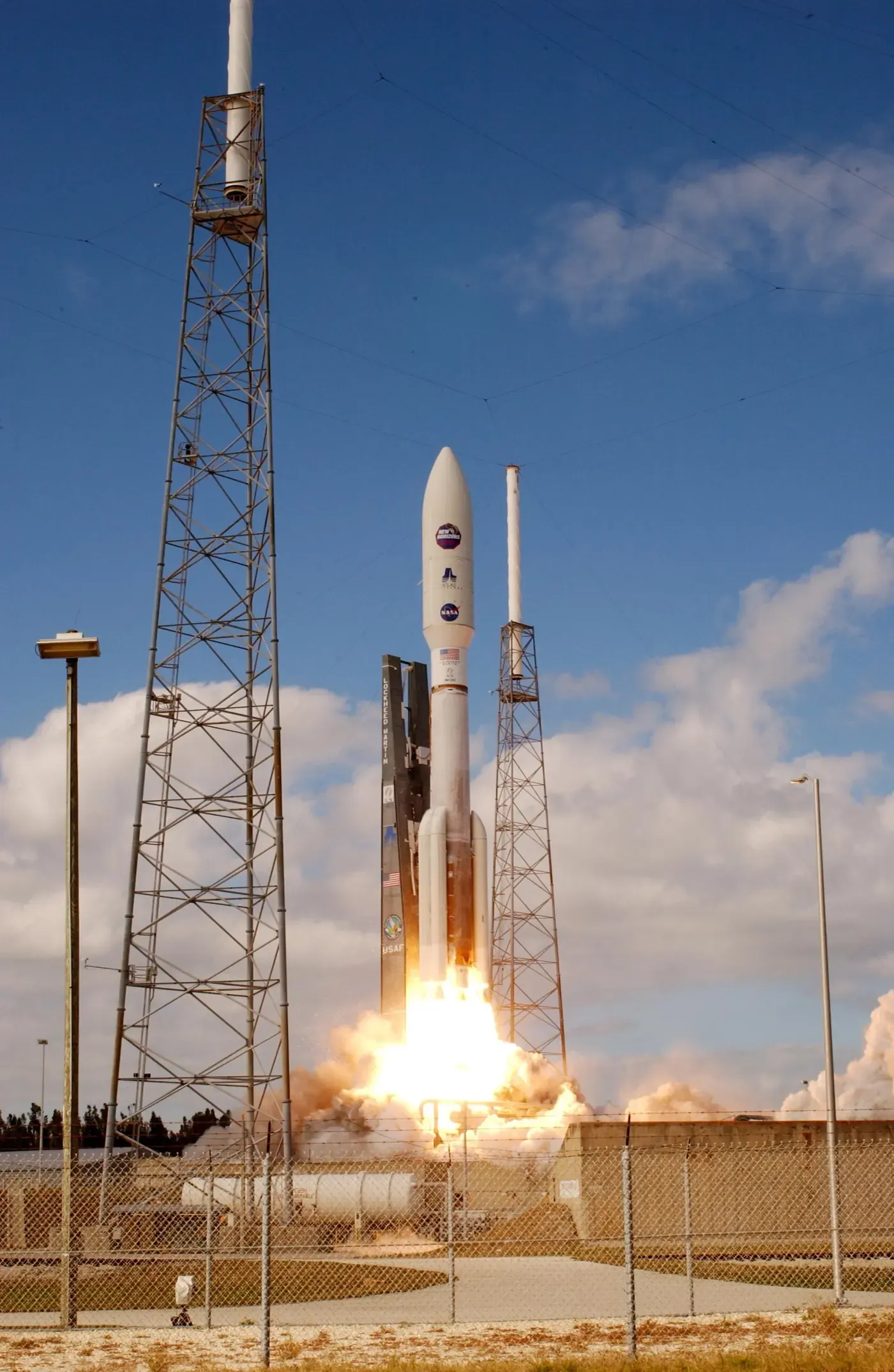 An Atlas V rocket blasting off from a launch pad in a burst of orange flame and white smoke, framed between two tall lightning-protection towers against a blue sky.