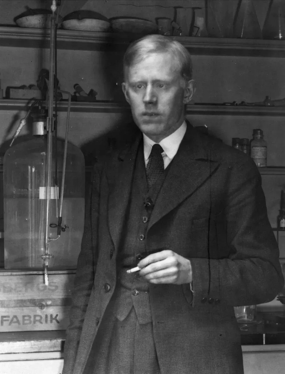 Odd Hassel standing in his laboratory in a three-piece suit, holding a cigarette, with glass apparatus and reagent bottles on the shelves behind him.