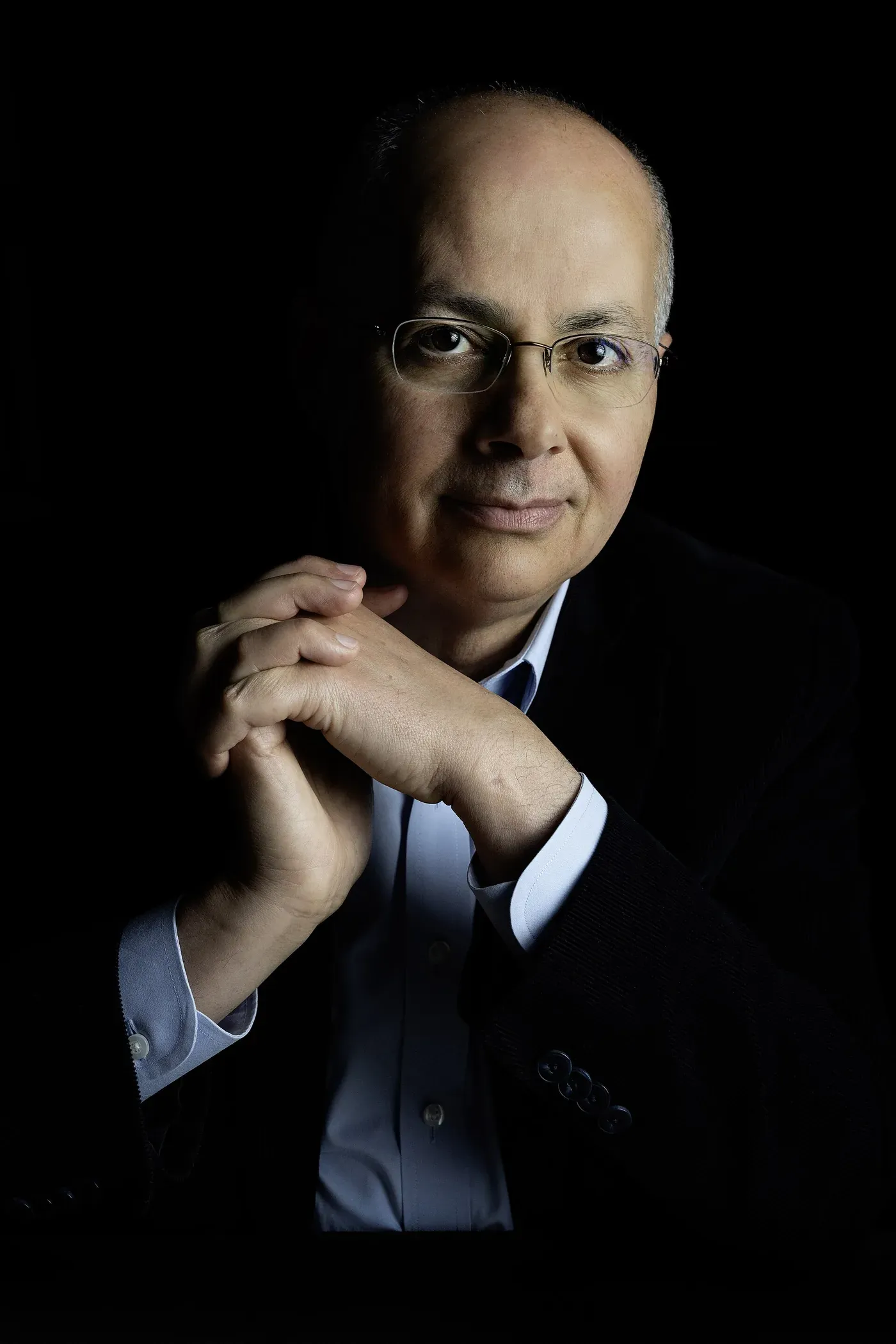 A close studio portrait, lit against a black background, of a balding man with glasses resting his chin on his clasped hands, wearing a dark jacket over a pale blue shirt.