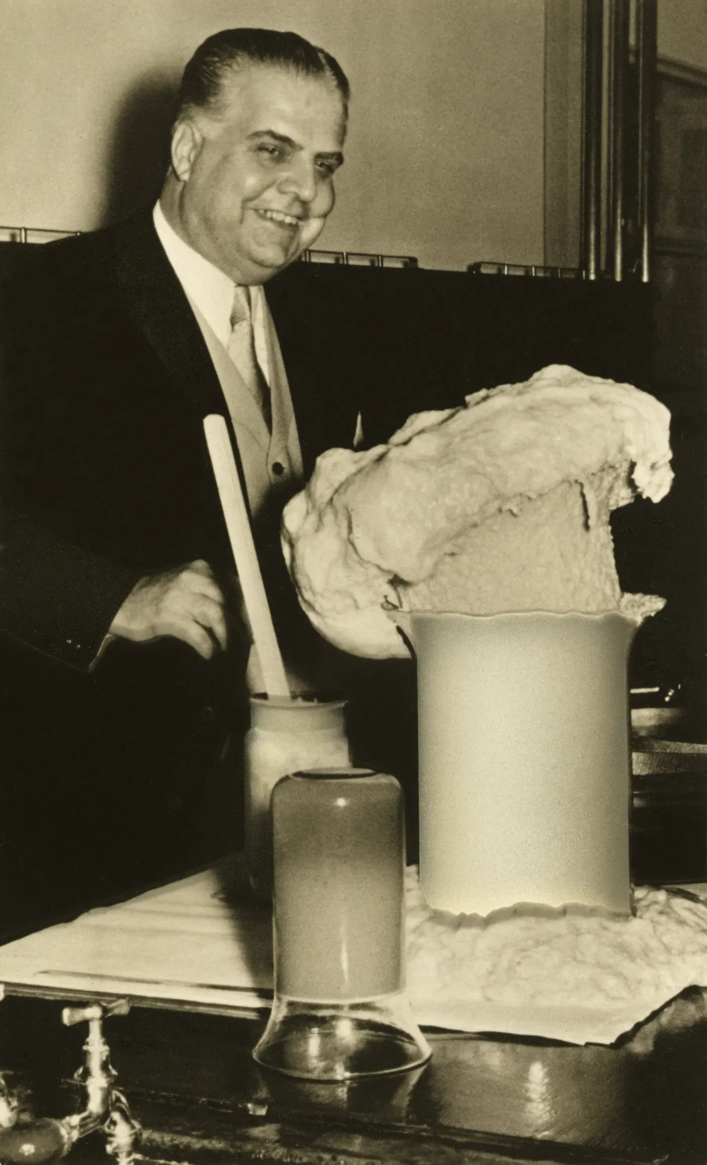 A smiling man in a suit stands over laboratory glassware from which a large mass of pale, irregularly bulging foam has risen and overflowed a beaker.