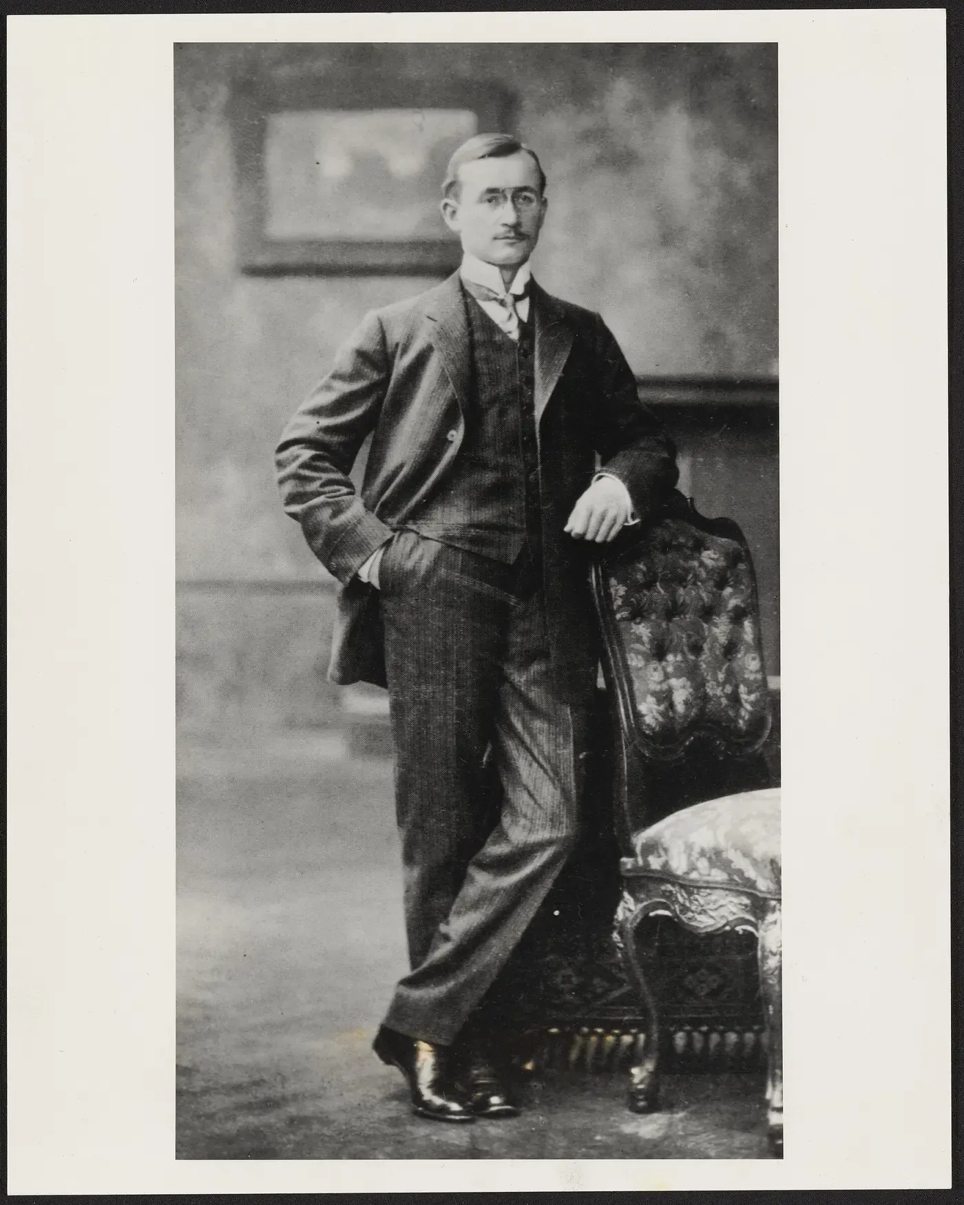 A formal studio portrait of a young man in a three-piece suit and tie, one hand in his trouser pocket, the other resting on the back of an ornately upholstered chair.