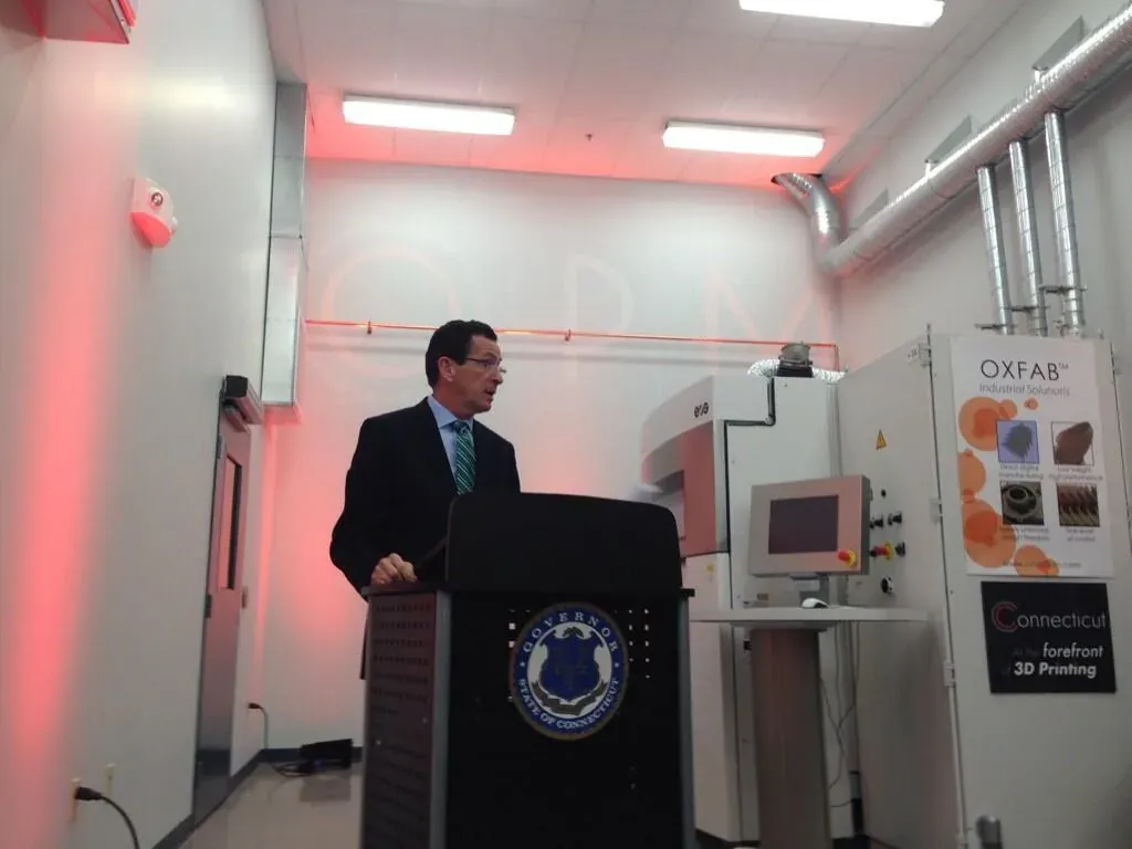 A man in a suit speaks from a lectern bearing the seal of the Governor of Connecticut, standing in front of a 3D-printing machine and a wall sign reading OXFAB Industrial Solutions.