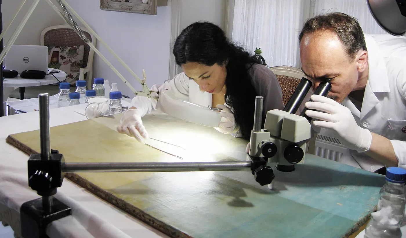 Two conservators in white coats and gloves examine a wooden panel painting through stereo microscopes and a magnifying lamp, surrounded by small labelled bottles on a workbench.