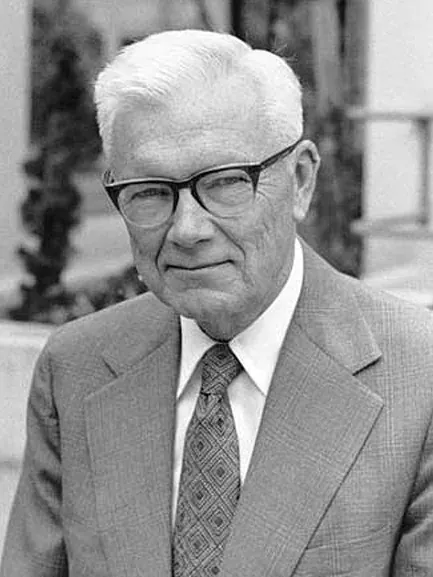 An elderly Paul Flory, in a tweed jacket and dark-framed glasses with a patterned tie, photographed outdoors.