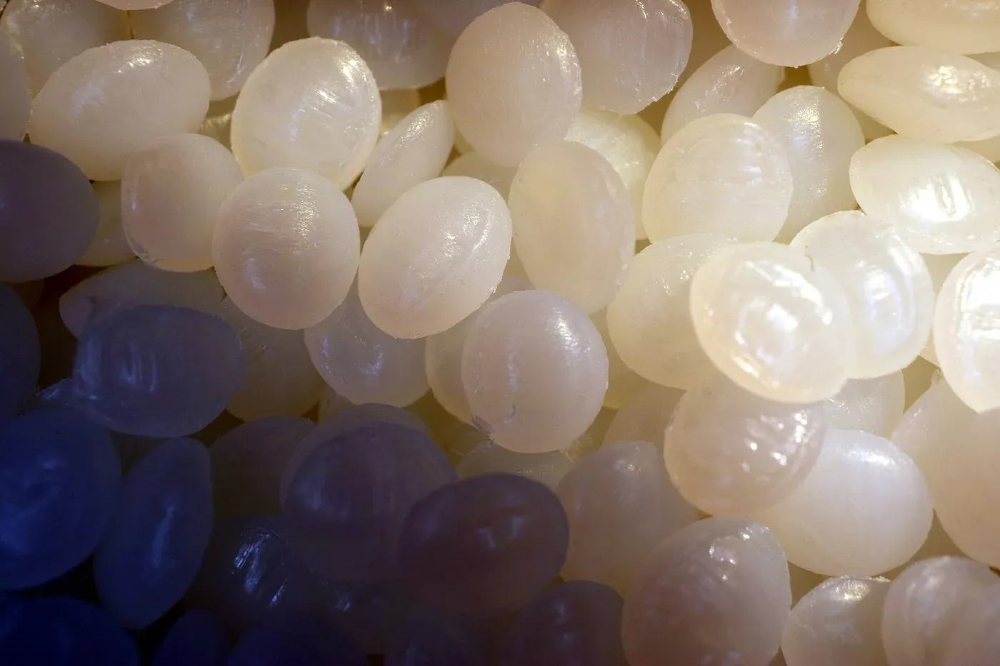 A close-up photograph of translucent, off-white plastic pellets, oval and pearl-like, densely packed together and catching the light unevenly.