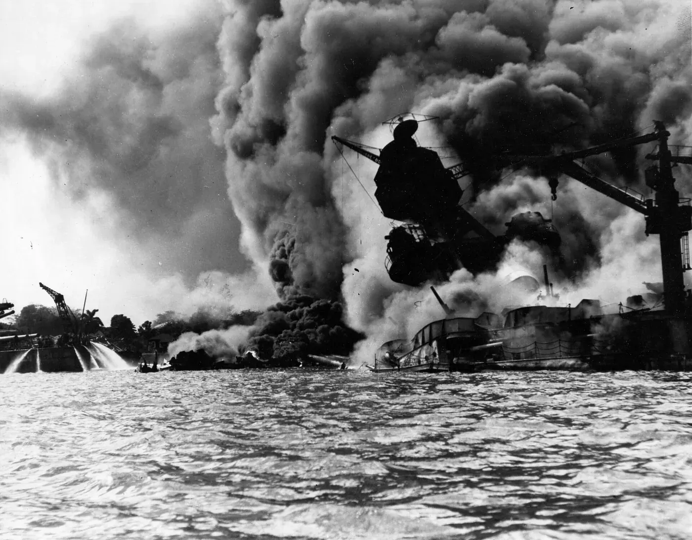 A warship listing and burning at anchor, engulfed in a vast column of black smoke, photographed from water level with a smaller vessel spraying firefighting water in the foreground.