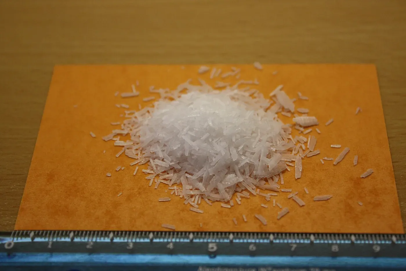 A small heap of translucent white waxy flakes on an orange card beside a ruler, showing the flakes span roughly five centimetres in width.