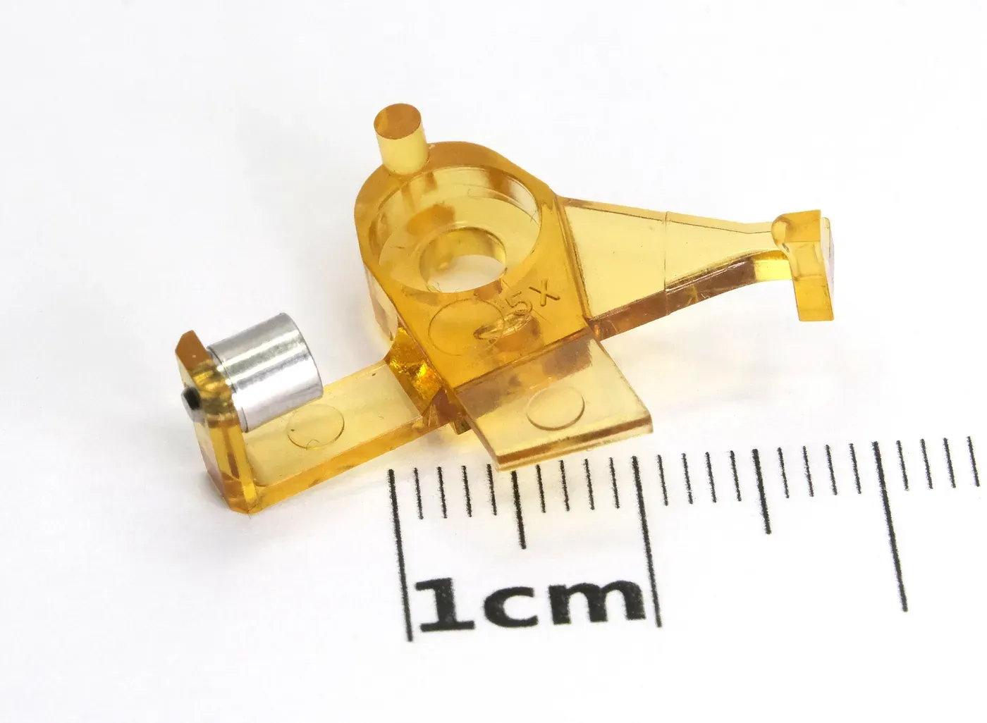 A small, transparent amber-coloured injection-moulded plastic bracket with a metal cylindrical insert, photographed on a white background next to a centimetre ruler.
