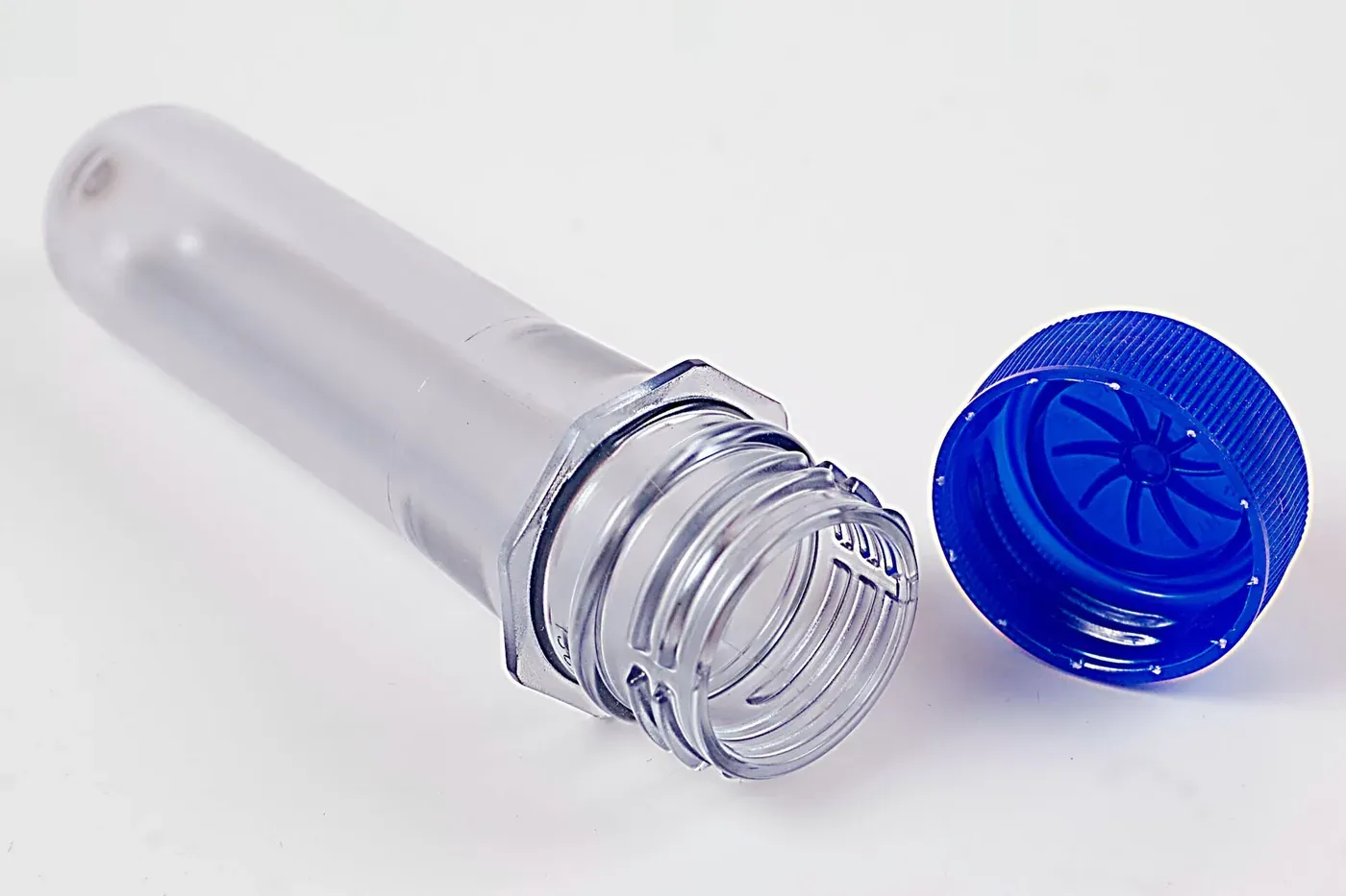 A clear, thick-walled PET preform shaped like a small test tube with a threaded neck, lying beside a separate blue plastic screw cap, on a white background.