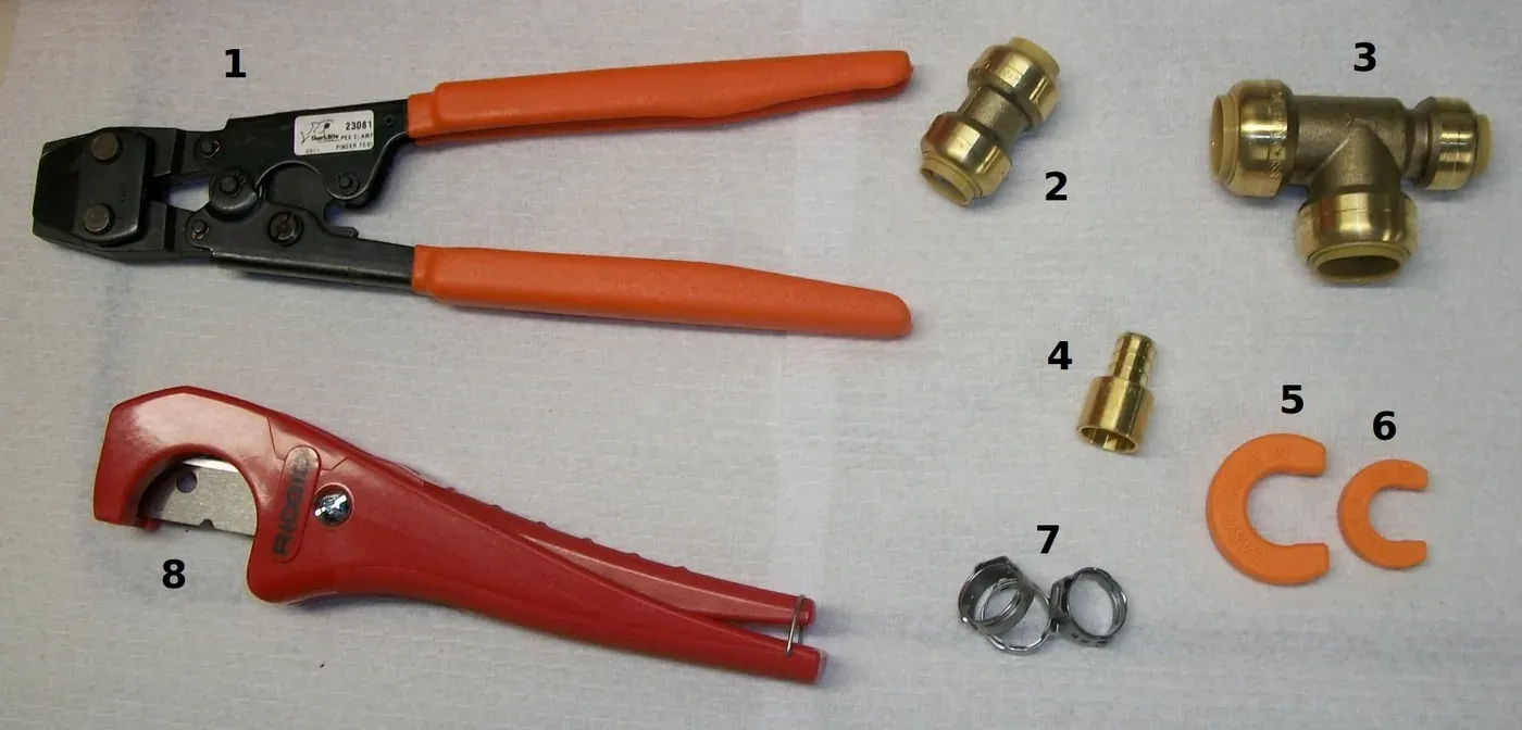 A flat-lay photograph of plumbing tools and brass fittings on a cloth surface: a crimping tool, a pipe cutter, push-fit couplings, a tee fitting and several stainless steel clamp rings.