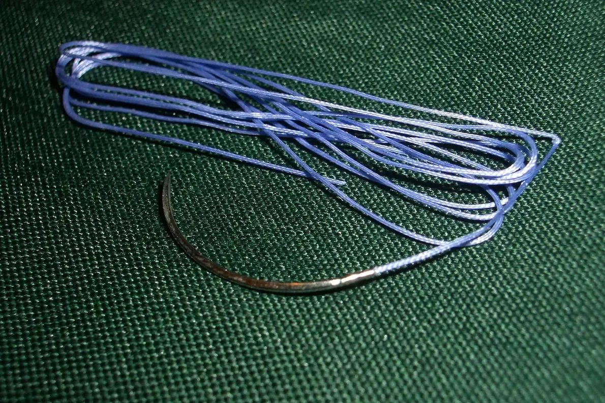 A pale blue braided suture thread coiled loosely on dark green cloth, with a curved metal needle swaged onto one end.