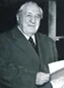 A small, low-resolution black-and-white photograph of an older man in a dark suit, seated indoors and holding a sheet of paper.