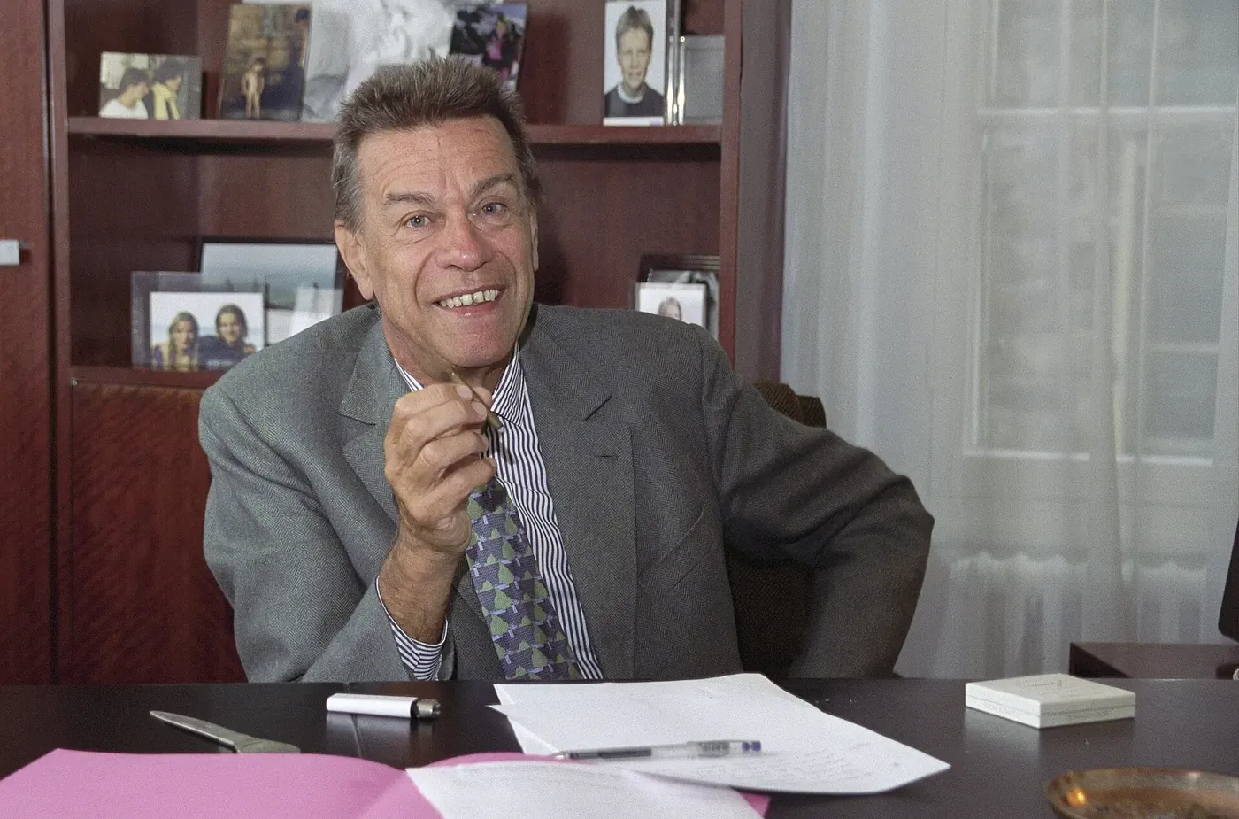 A man in a grey jacket and patterned tie sitting at a desk in an office, gesturing with a pen in hand and smiling, with framed photographs on a shelf behind him.