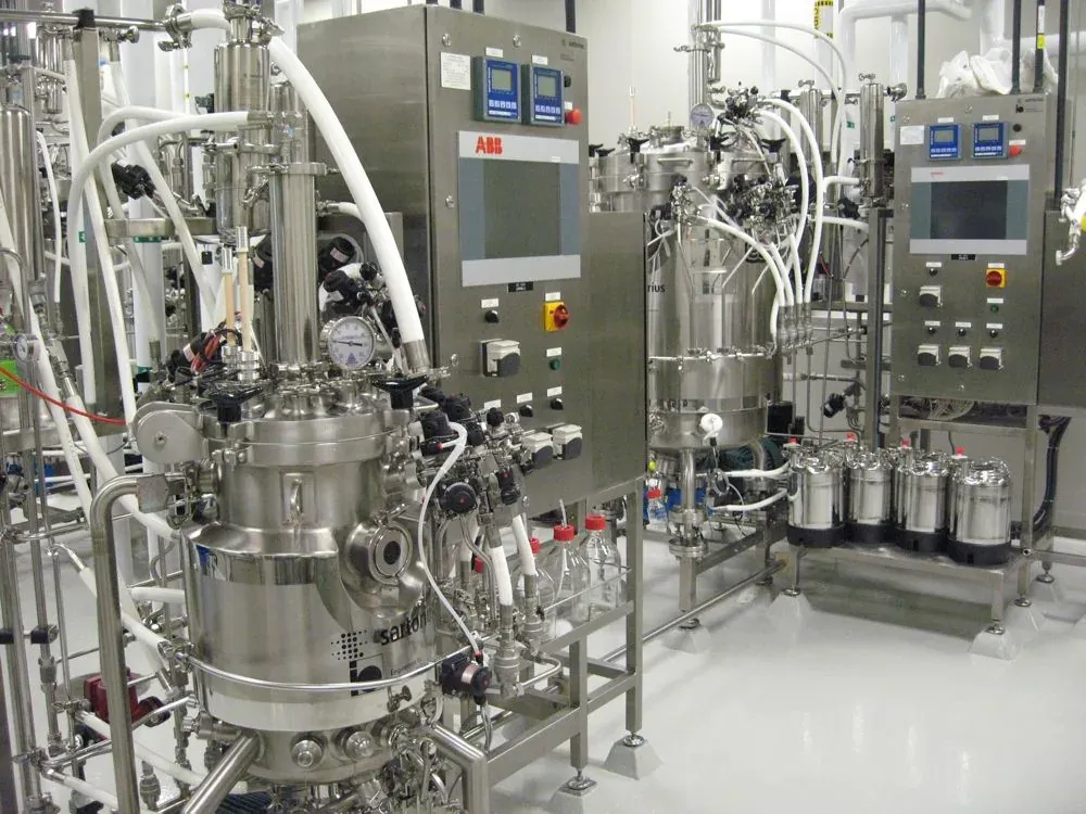 A row of polished stainless-steel bioreactor vessels with control panels, sensors, and coiled tubing, in a clean industrial laboratory setting.