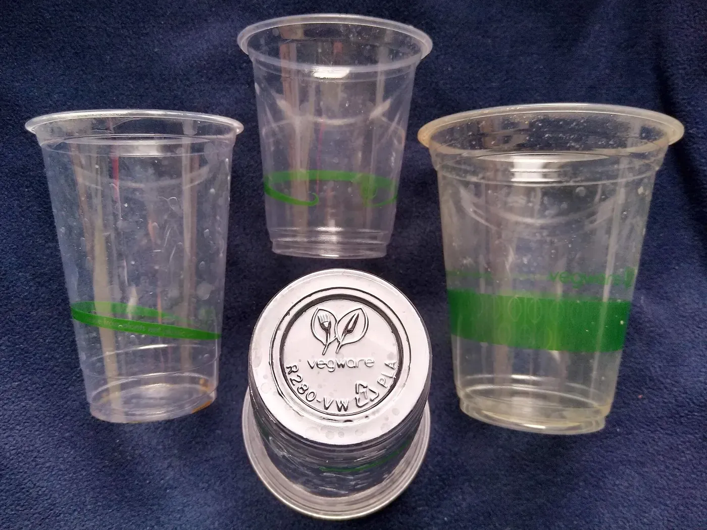 Four clear plastic cups standing on a dark cloth, each printed with a green band and leaf logo, with one cup's base turned upward to show a stamped recycling symbol reading VEGWARE R280-VW PLA.
