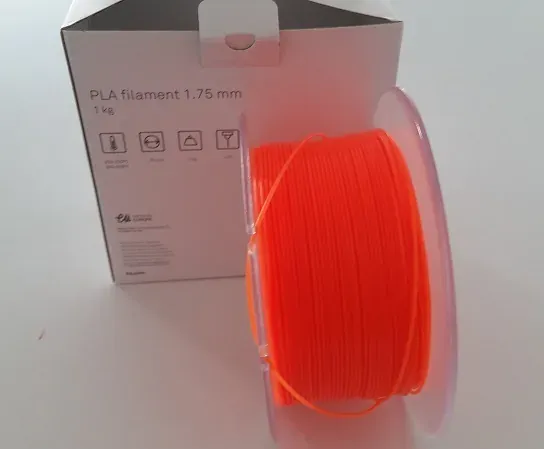 A spool of bright orange plastic filament wound in front of its retail box, the box printed with the text 'PLA filament 1.75 mm, 1 kg' and small icons for storage and printing conditions.