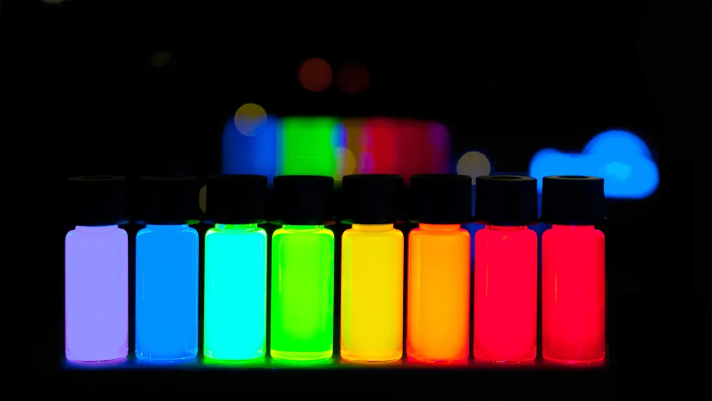 Eight small glass vials in a row, each glowing a different vivid colour under ultraviolet light (from pale violet through cyan, green, yellow, orange, and two shades of red) against a black background.