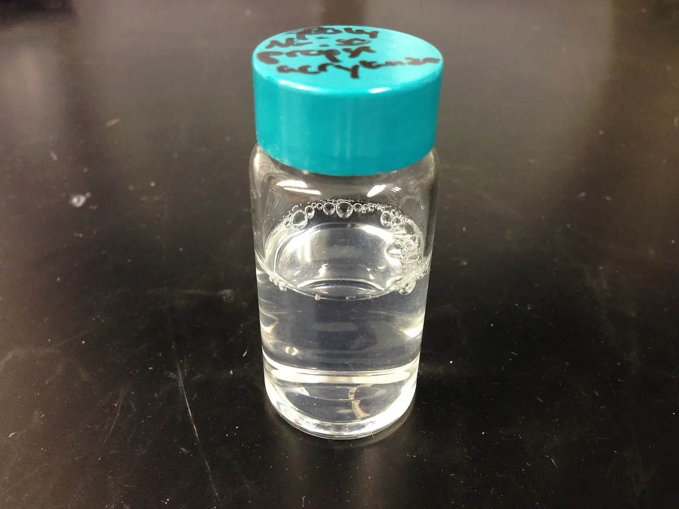 A small glass sample vial with a teal cap, containing a clear, colorless liquid at room temperature.