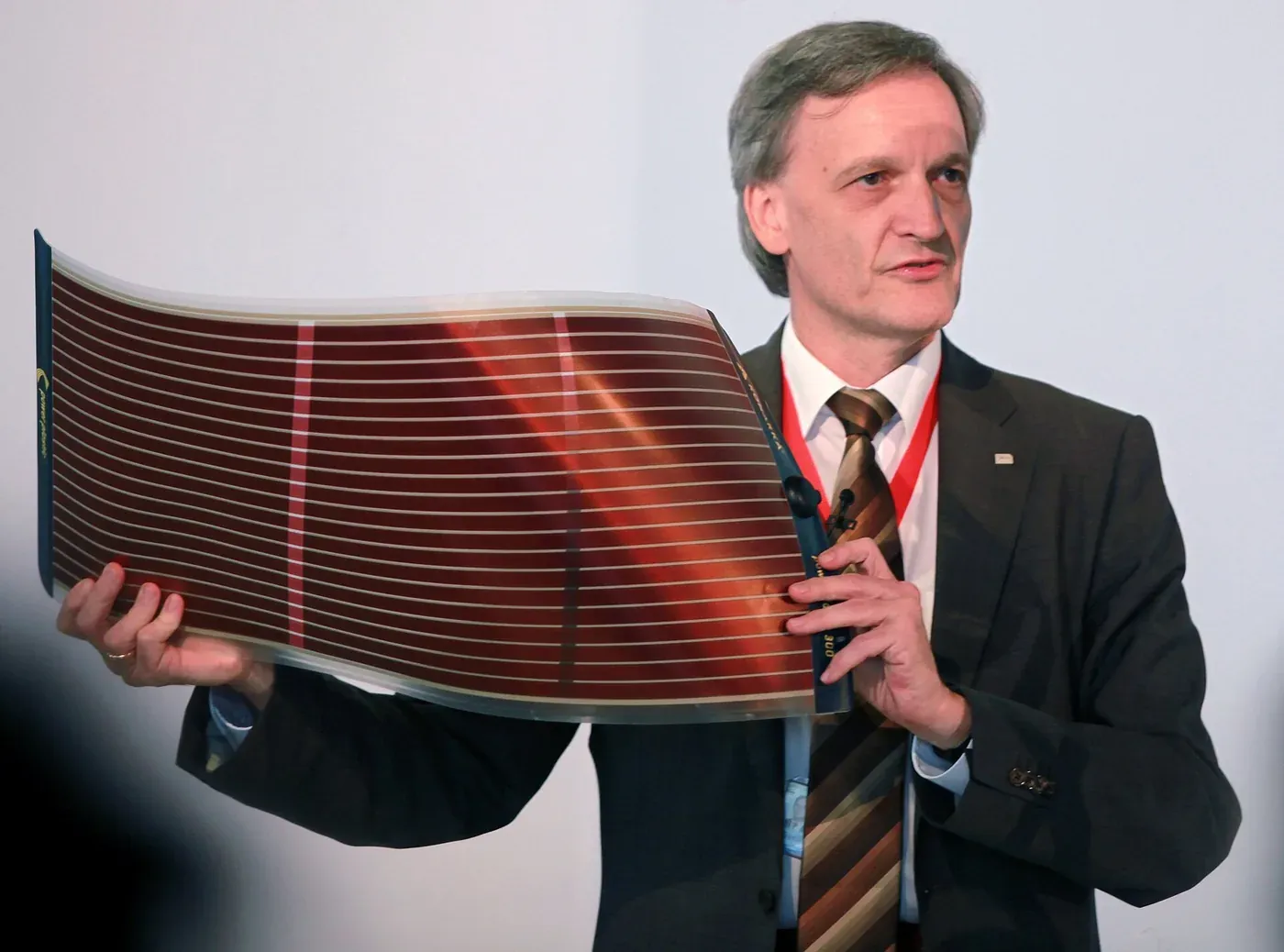 A man in a suit holds up and flexes a large, flexible reddish-brown striped solar panel sheet at a press event.