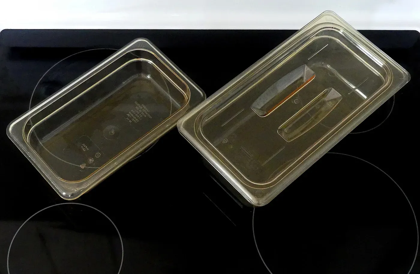 Two amber-tinted transparent plastic food pans, one holding a slotted spoon and a pair of tongs, sitting on a black glass cooktop.