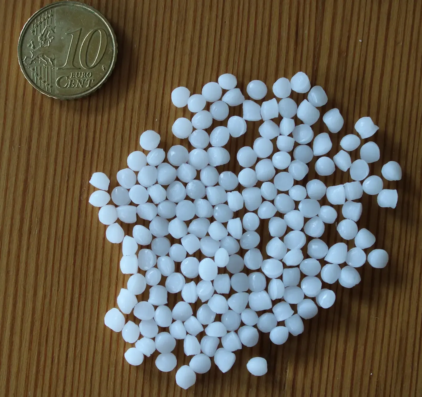 A scattered pile of small white plastic pellets on a wood-grain surface, shown next to a euro coin for scale.