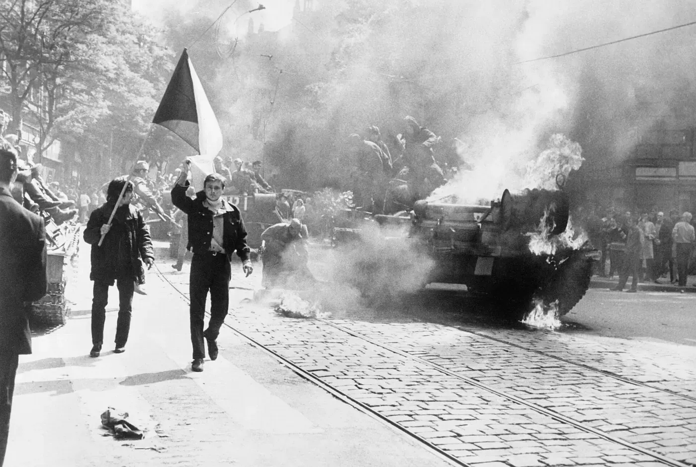 Two young men walk along a cobbled Prague street past a burning tank wreathed in smoke, one of them carrying a Czechoslovak flag, as onlookers watch from the pavement and soldiers stand atop a second armoured vehicle behind it.
