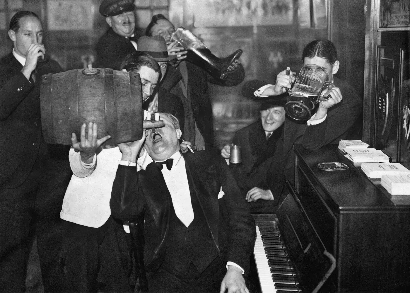 Black-and-white photograph of men in formal evening dress crowded around a piano, drinking beer straight from a keg and from large glasses and mugs.