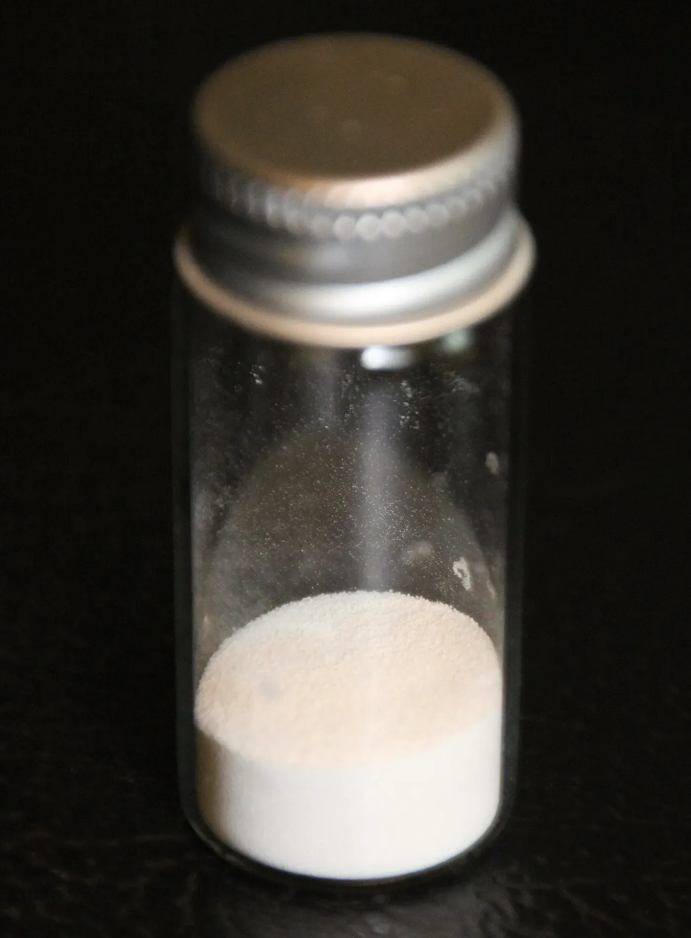 A small glass vial, capped with a metal screw lid, half filled with a fine white powder.