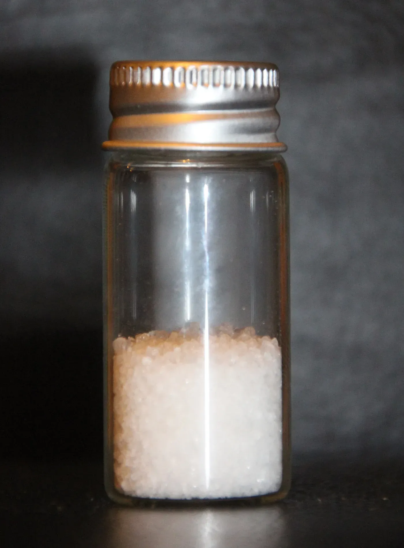 A small glass vial holding pale, granular solid pellets of polyvinyl alcohol, photographed against a dark background.