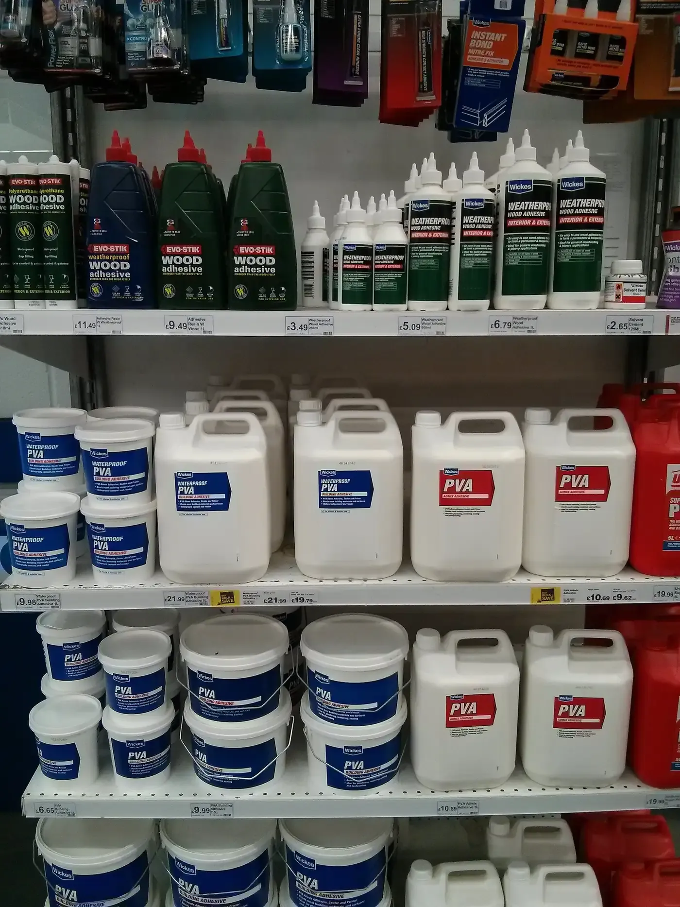 A hardware-store shelf stocked with dozens of bottles, tubs and jerrycans of PVA wood adhesive and building adhesive under various brand names.