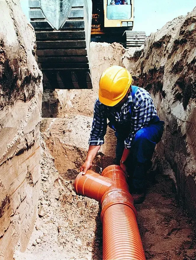 A construction worker in a hard hat kneels in a trench, fitting together large orange-brown ribbed plastic pipe sections, with an excavator bucket visible above.