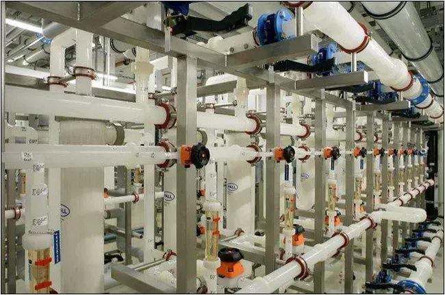 An indoor industrial installation of white plastic piping racks with numerous blue and orange valves, flow meters and fittings, arranged in parallel rows connected by overhead pipework.