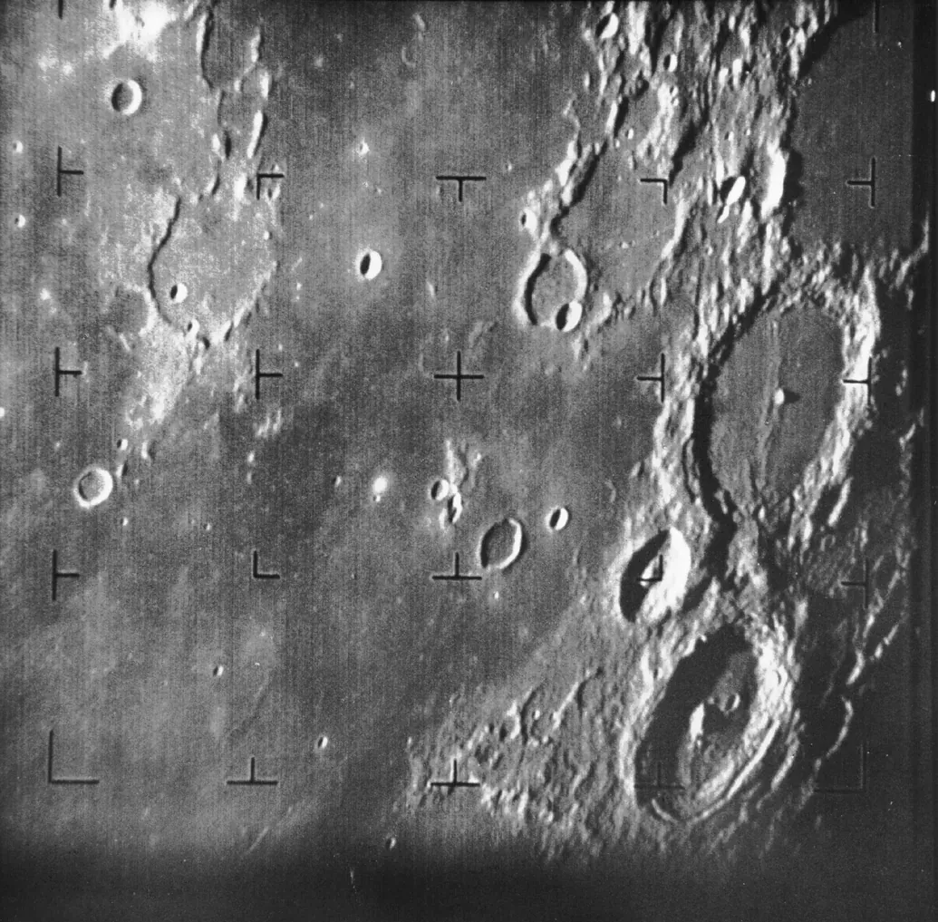A grainy black-and-white photograph of the Moon's cratered surface, densely pocked with craters of many sizes, with small crosshair calibration marks along the frame's edges.